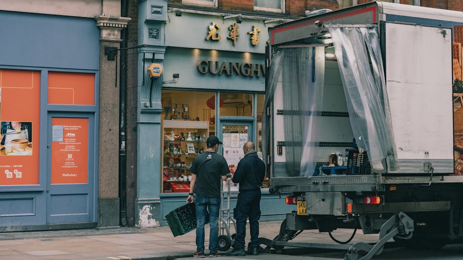 A silver flatbed van used for house removals is captured in motion on an urban street during evening or night time, with a blurred background of illuminated buildings and a pinkish sky. The van is pos