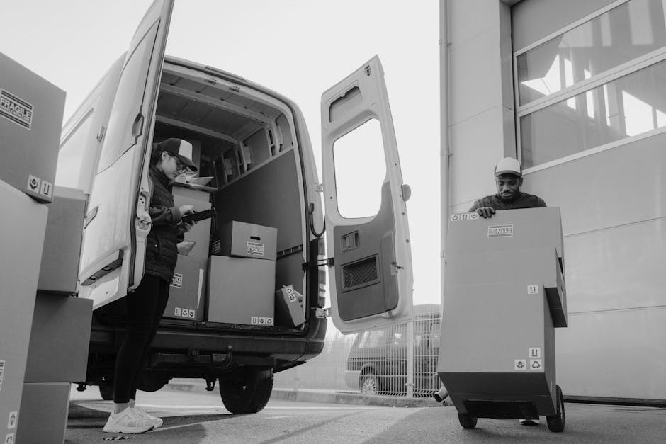 A vintage cream-colored van is shown in motion on a highway, with a blurred background indicating speed. The vehicle's flatbed cargo area is loaded with various household furniture items, including wo
