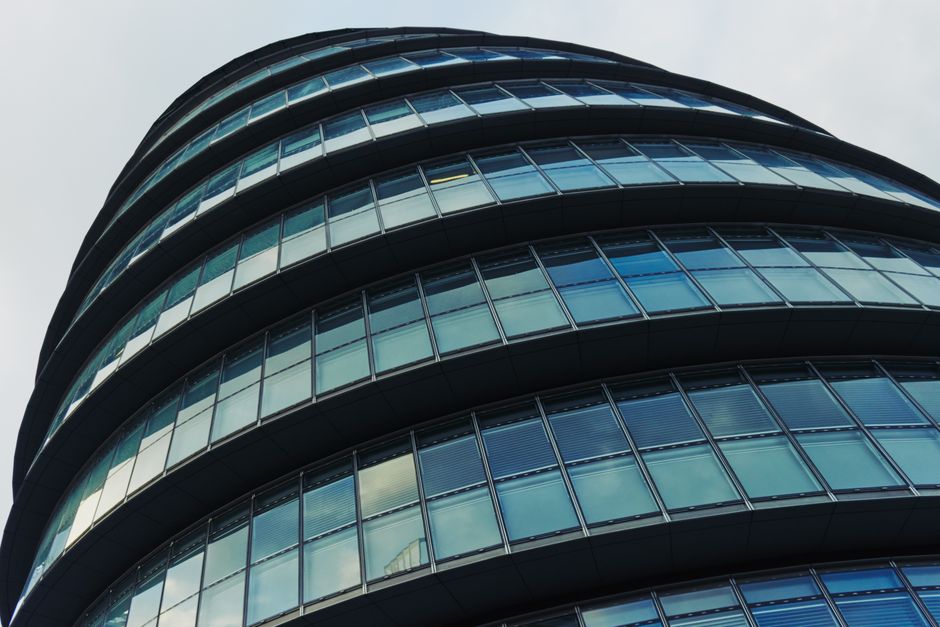 A modern multi-storey office building with a curved facade, featuring multiple floors of glass windows framed by black metal. The glass panels reflect the cloudy sky above, and the building's rounded