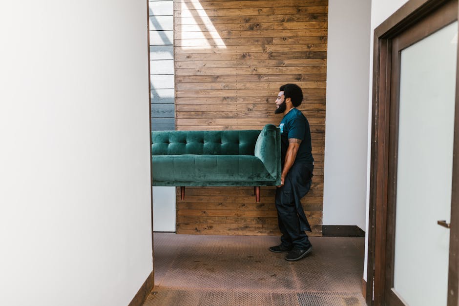 A man with dark curly hair and a beard is inside a bright, minimalist room with white walls and large arched windows in the background, natural light illuminating the space. He is wearing a dark blue